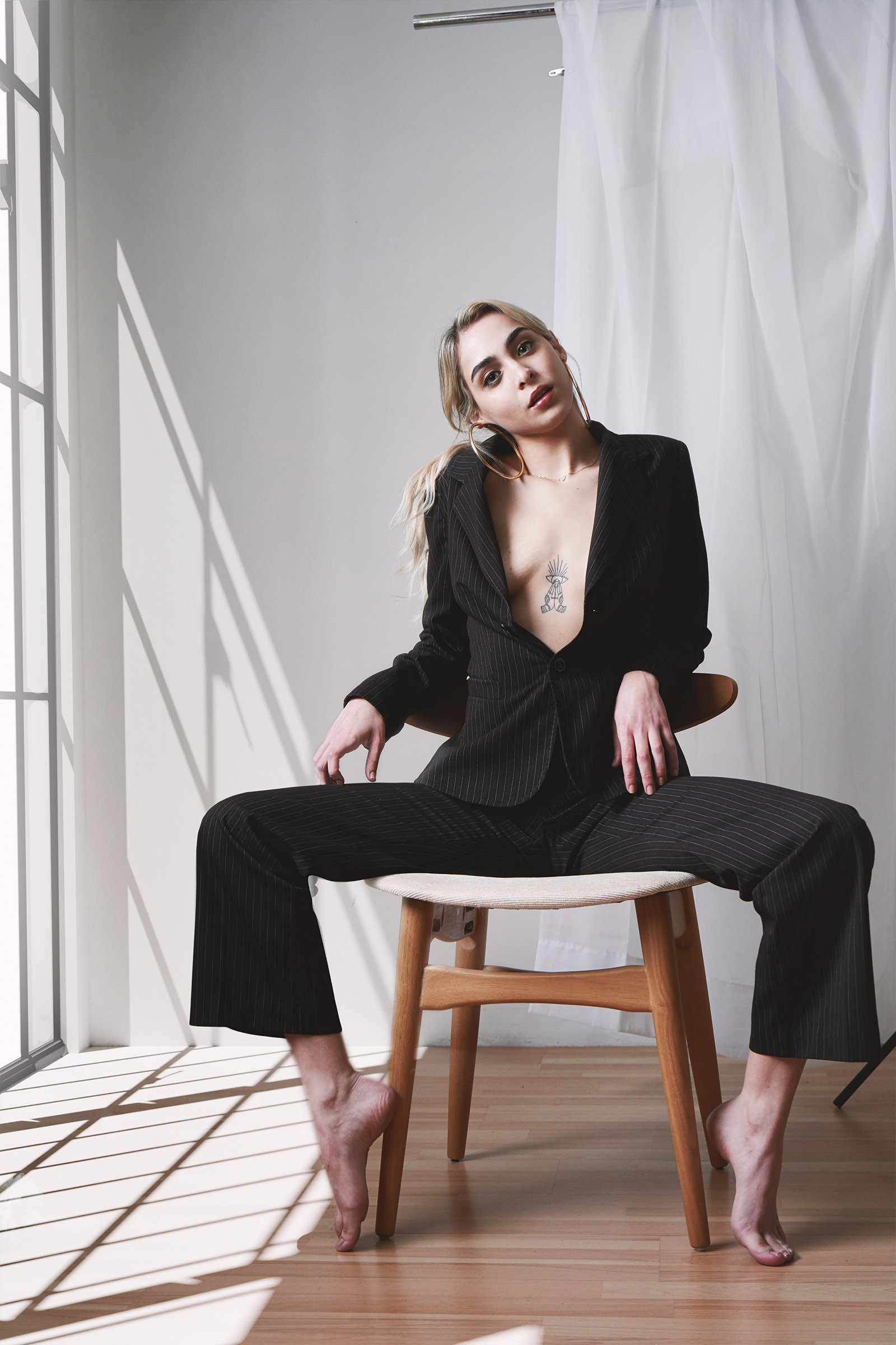 Fernanda Padilla sits elegantly on a wooden chair wearing a black pinstriped suit with a tattoo on her chest against a backdrop of a white curtain and sunlit room.
